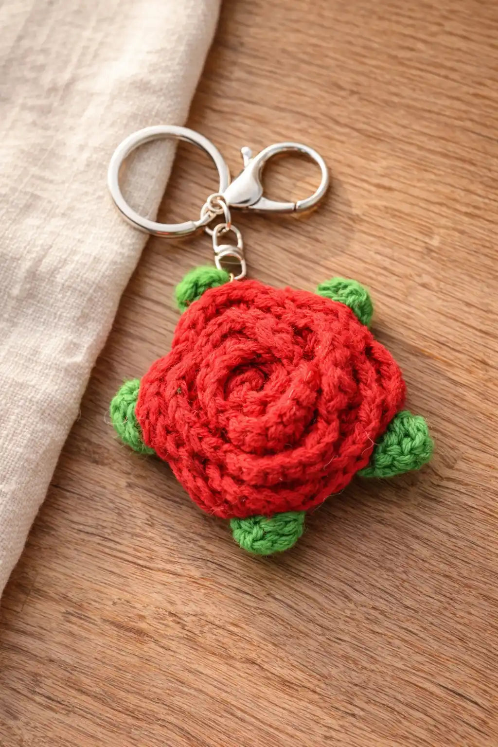 Detailed view of a red crochet rose keychain with green leaves and a silver lobster clasp on a wooden surface.