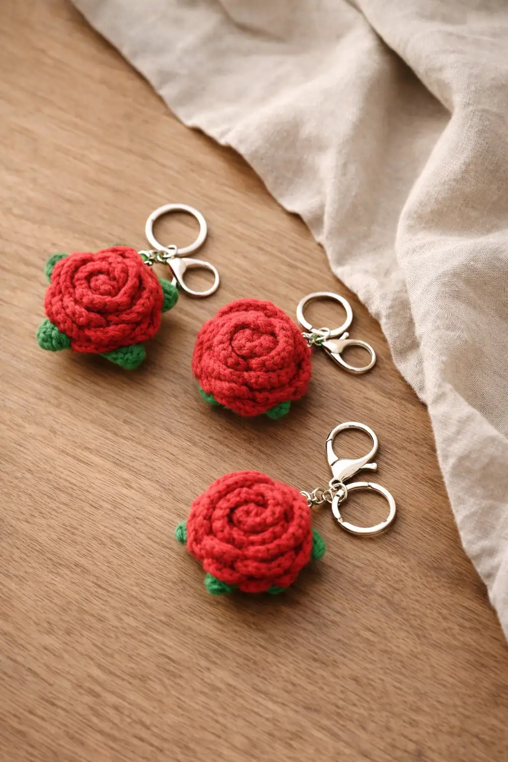 Detailed view of a red crochet rose keychain with green leaves and a silver lobster clasp on a wooden surface.